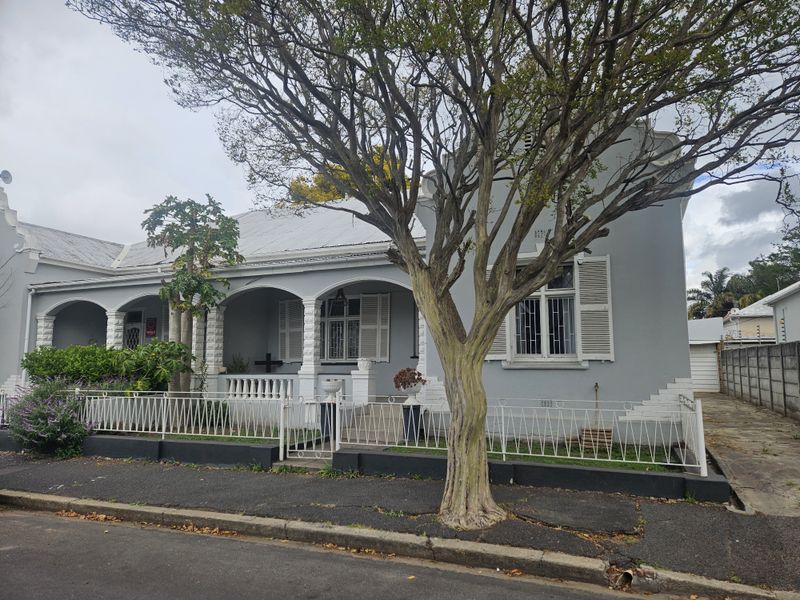 Charming historic-style house featuring a covered front veranda, decorative columns, and secure metal fencing, set behind a mature tree.