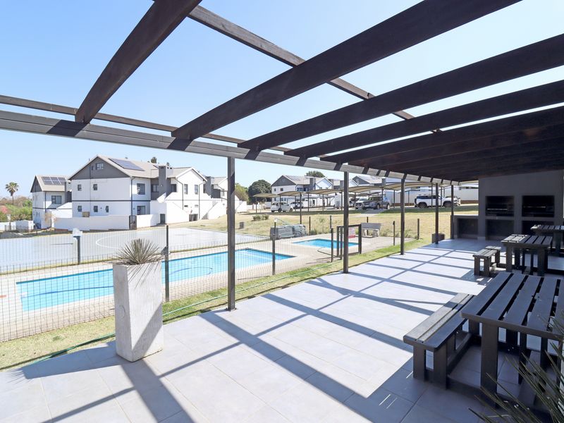 Covered outdoor patio featuring a timber pergola, built-in braai, and tiled flooring, overlooking a communal swimming pool and residential complex grounds.