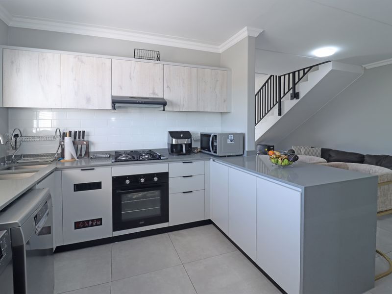 Modern open-plan kitchen featuring light wood-look upper cabinets, stone countertops, gas hob, and integrated oven, seamlessly flowing into the living area with staircase access.