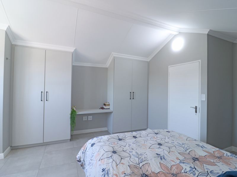 A modern bedroom featuring neutral grey walls, built-in cupboards with integrated desk space, tiled flooring, and a vaulted ceiling with coving.