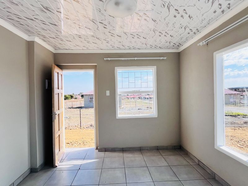 A bright, empty living area featuring tiled floors, decorative ceiling panels, and security bars on the windows, with direct access to the exterior.