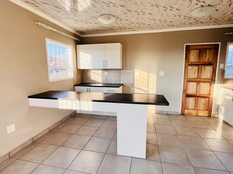 A bright, open-plan kitchen featuring modern white cabinetry, dark laminate countertops, a breakfast bar, and neutral tiled flooring.