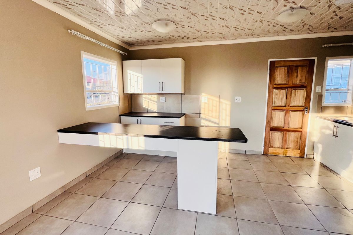A bright, open-plan kitchen featuring modern white cabinetry, dark laminate countertops, a breakfast bar, and neutral tiled flooring.