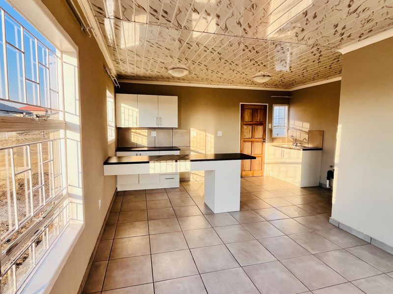 An open-plan kitchen and living area featuring tiled floors, modern cabinetry, a central island, and decorative ceiling panels, bathed in natural light.