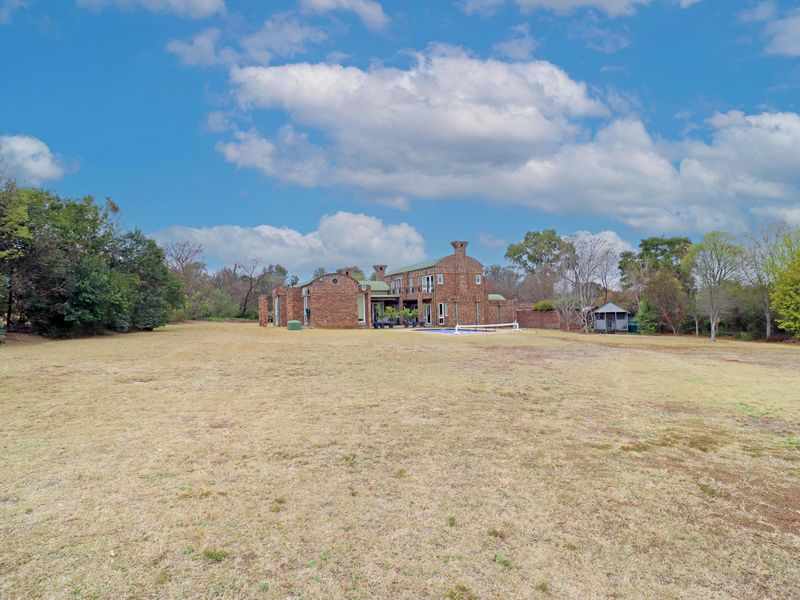 A wide-angle view of the rear of the brick house, featuring a large lawn, mature landscaping, and a swimming pool.