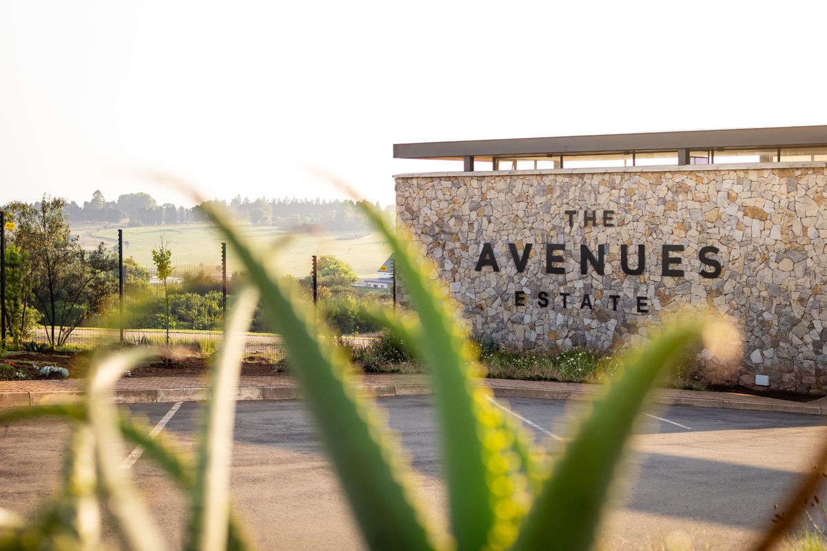 The entrance to The Avenues Estate featuring a prominent stone-clad wall, paved driveway, and secure perimeter fencing.