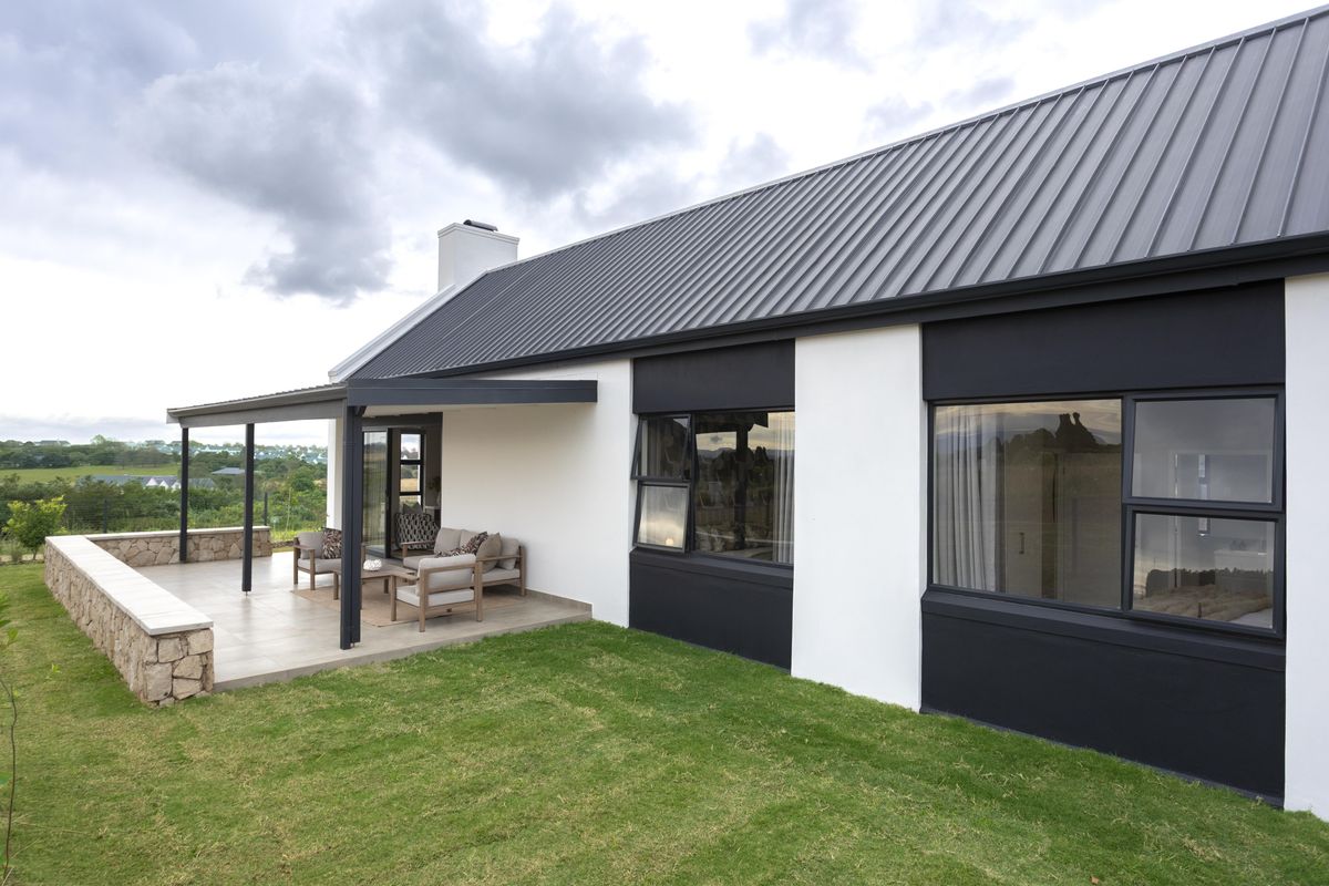 A modern rear exterior view featuring a covered patio with stone retaining wall, metal roof, and large windows overlooking the garden.