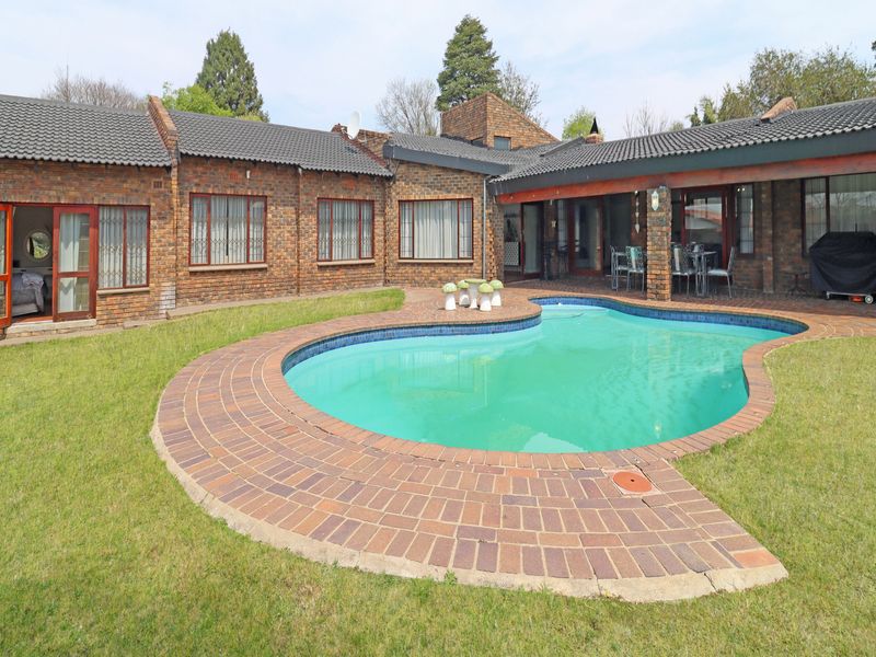 A rear garden featuring a kidney-shaped swimming pool with brick paving, a covered patio area, and a face brick house exterior.