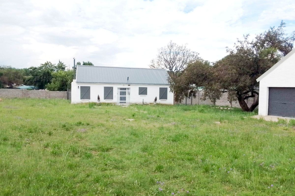 Rear view of the property featuring a single-story dwelling with a metal roof, security shutters, and a detached garage set on a large grassed plot.