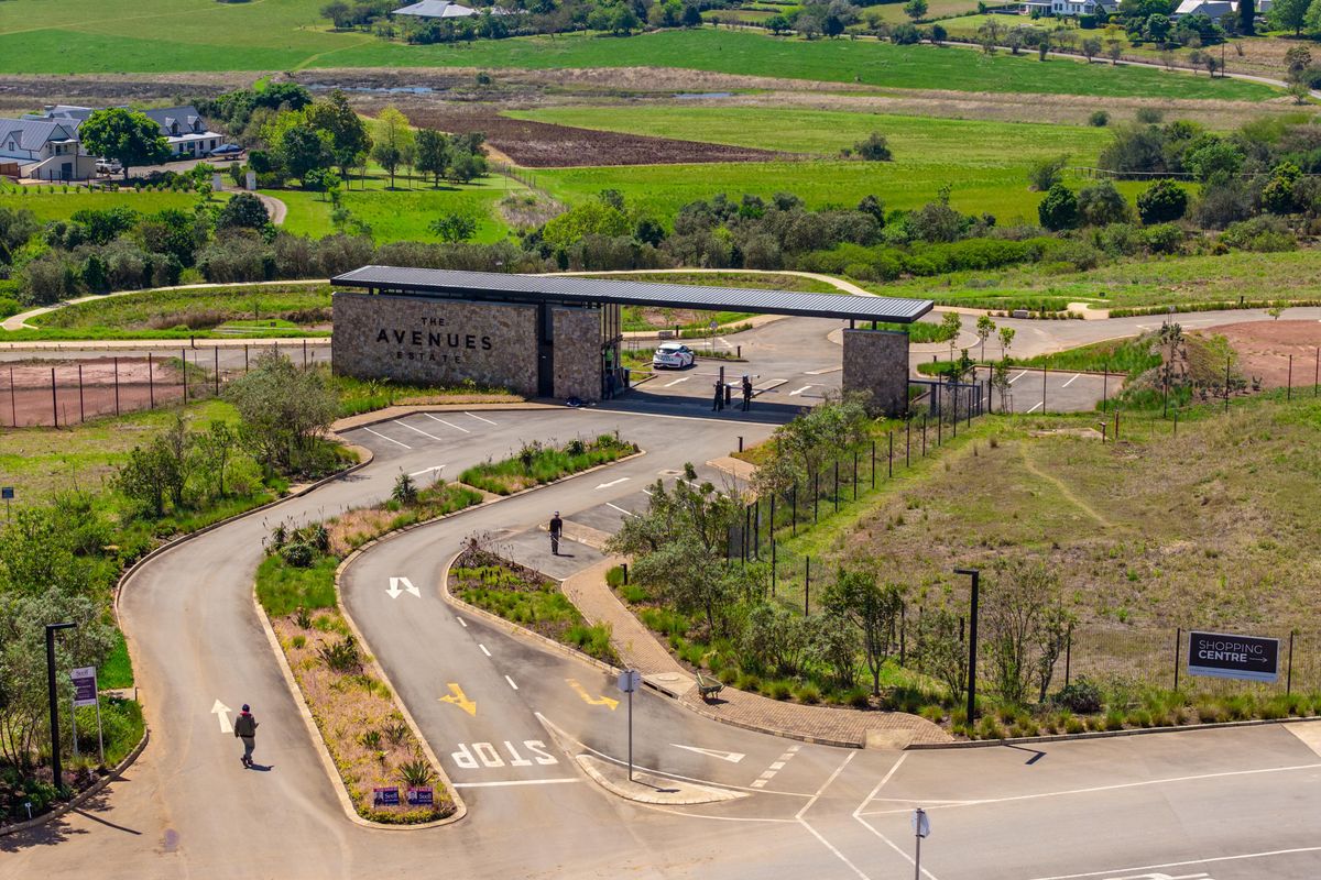 Aerial view of The Avenues Estate entrance, featuring a modern stone-clad security gatehouse, paved access roads, and landscaped surroundings.