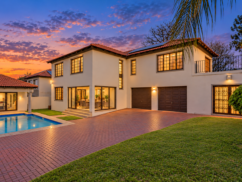 A beautifully maintained double-story home featuring a swimming pool, paved driveway, double garage, and a well-manicured garden under a vibrant sunset sky.