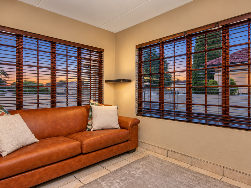 A bright living area featuring large windows with wooden venetian blinds and neutral tiled flooring, offering a comfortable and well-lit space.