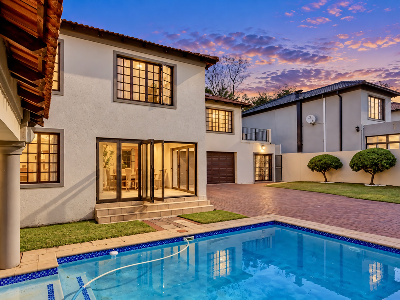 A well-maintained rear exterior view featuring a swimming pool, paved driveway, covered patio area, and bi-fold doors leading into the home.