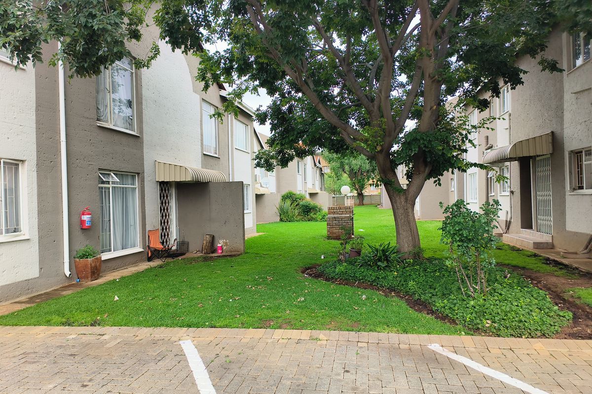 A view of the apartment complex exterior featuring a shared green lawn, mature trees, and paved parking area.