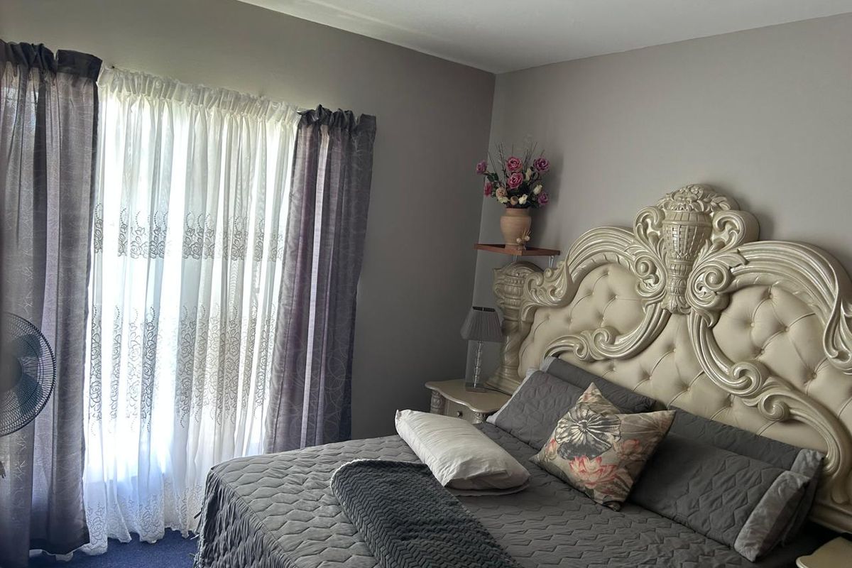 A bedroom featuring neutral wall tones, carpeted flooring, and large windows with layered curtains.