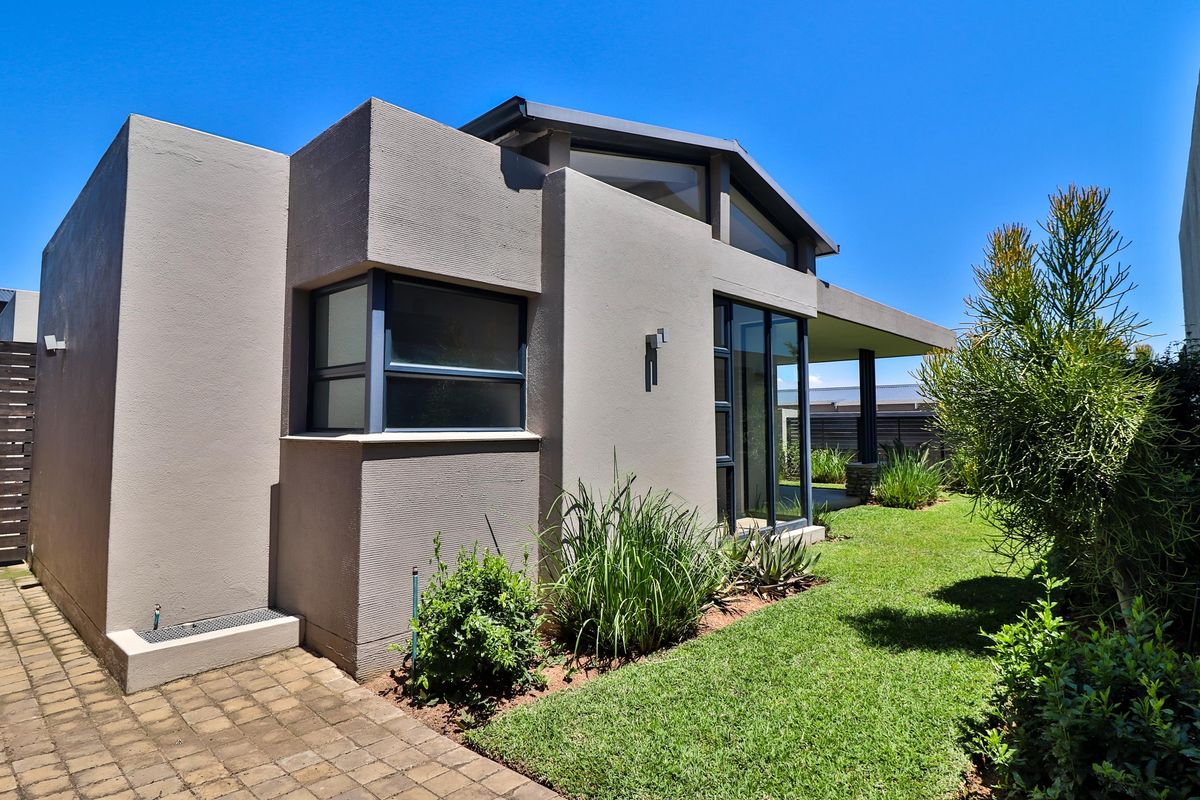 A modern rear exterior view of the house featuring a well-maintained garden, paved walkway, and a covered patio area with contemporary architectural lines.