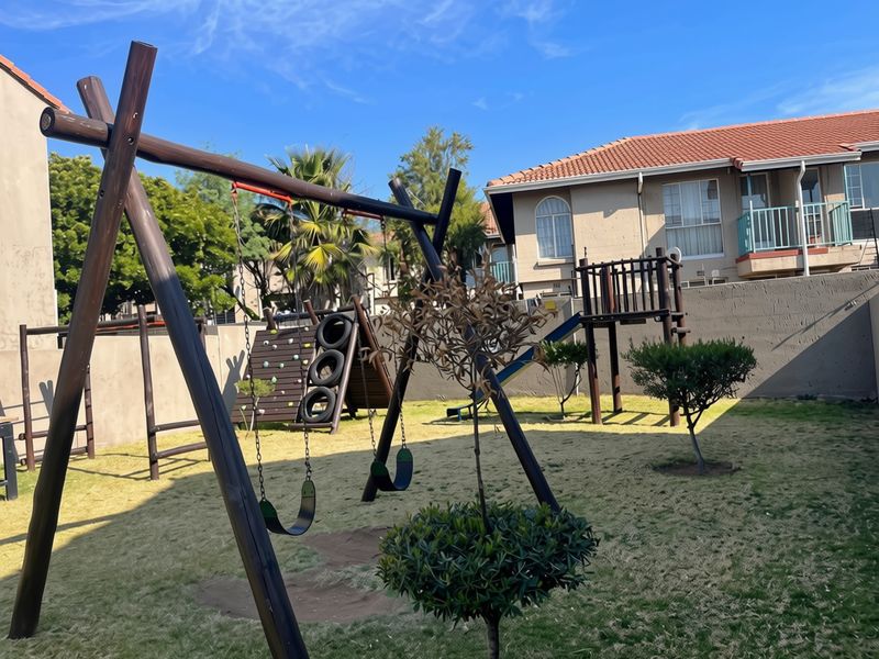 A communal garden area featuring a wooden playground structure with swings, a climbing wall, and a slide, set against a residential apartment building.
