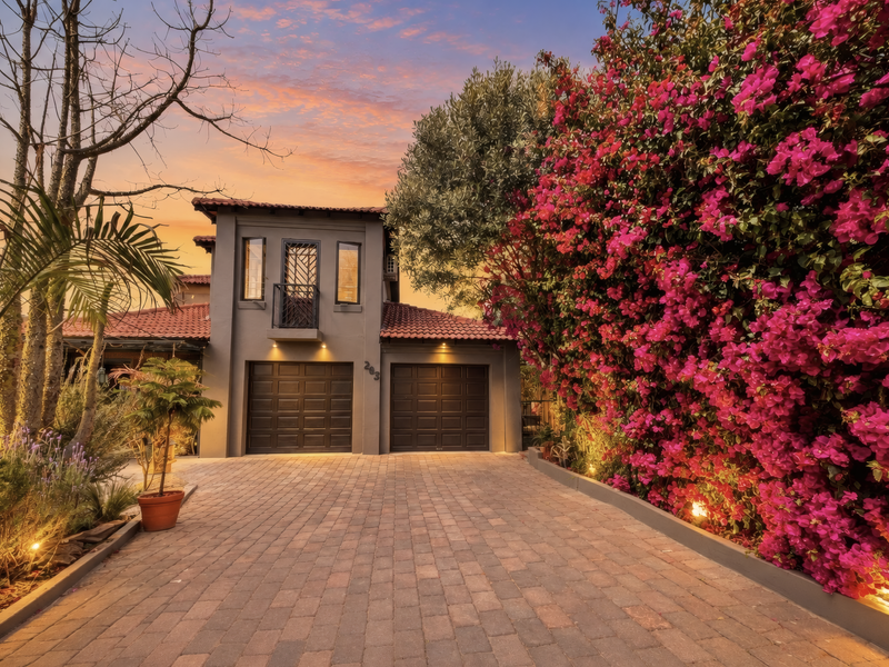 A well-maintained townhouse exterior featuring a double garage, paved driveway, and a charming balcony, complemented by vibrant bougainvillea and sunset lighting.