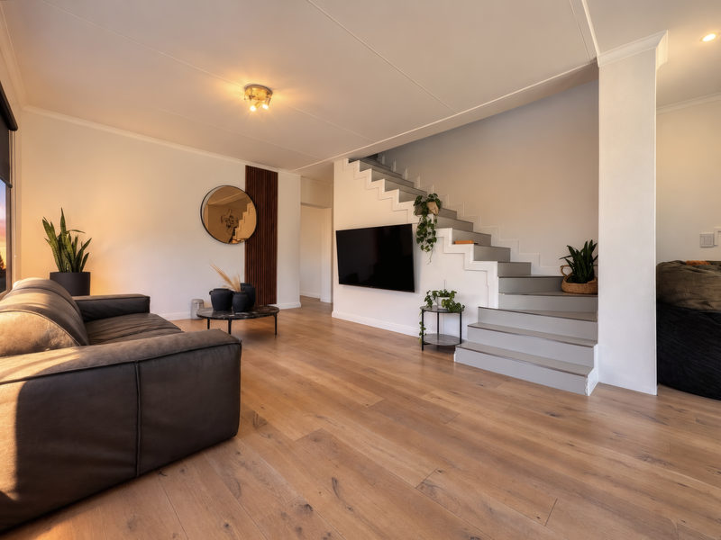 A bright and modern living room featuring light wood-look laminate flooring, a sleek staircase, and a wall-mounted television.