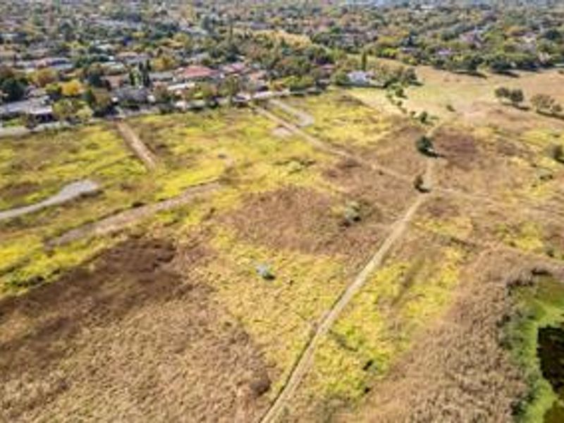 An aerial view of a large, undeveloped residential land plot featuring natural vegetation and dirt access paths, situated near an established suburban neighborhood.