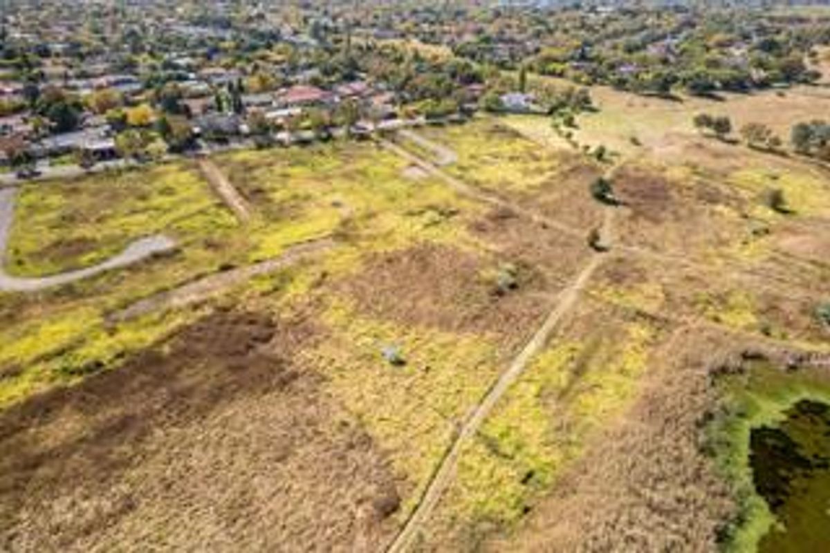 An aerial view of a large, undeveloped residential land plot featuring natural vegetation and dirt access paths, situated near an established suburban neighborhood.
