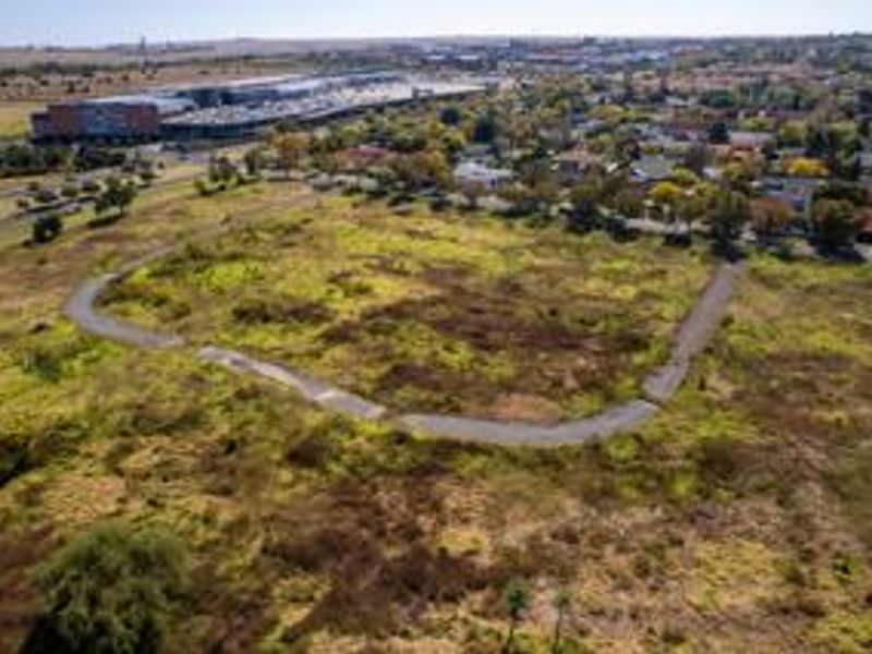 Aerial view of a large vacant residential plot featuring natural vegetation and a paved access path, situated near established suburban infrastructure.