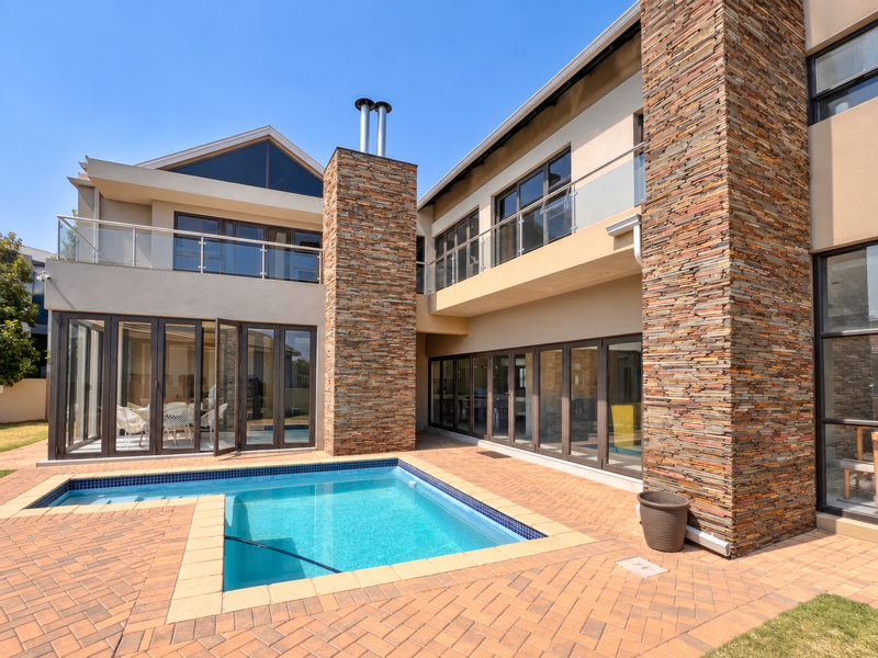 Modern double-story residence featuring stone cladding, expansive glass bi-fold doors, glass-railed balconies, and a private swimming pool set within a paved patio area.