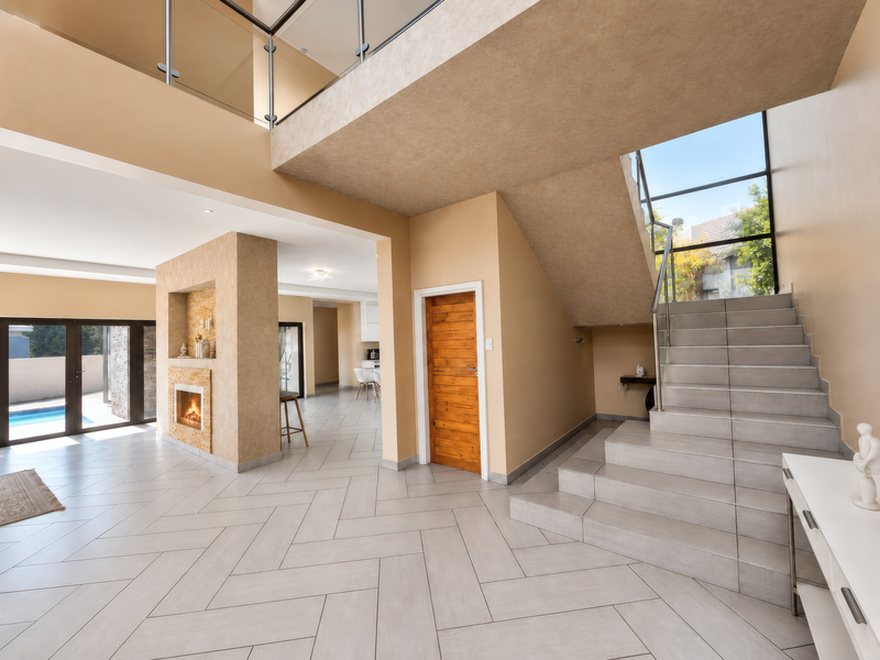 Spacious open-plan living area featuring a double-volume ceiling, modern tiled flooring, a built-in fireplace, and a striking staircase with a glass balustrade.