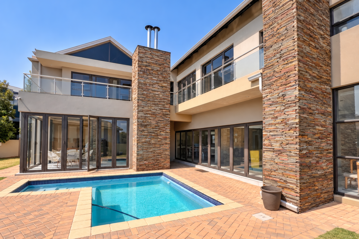 Modern double-story residence featuring stone cladding, expansive glass bi-fold doors, glass-railed balconies, and a private swimming pool set within a paved patio area.