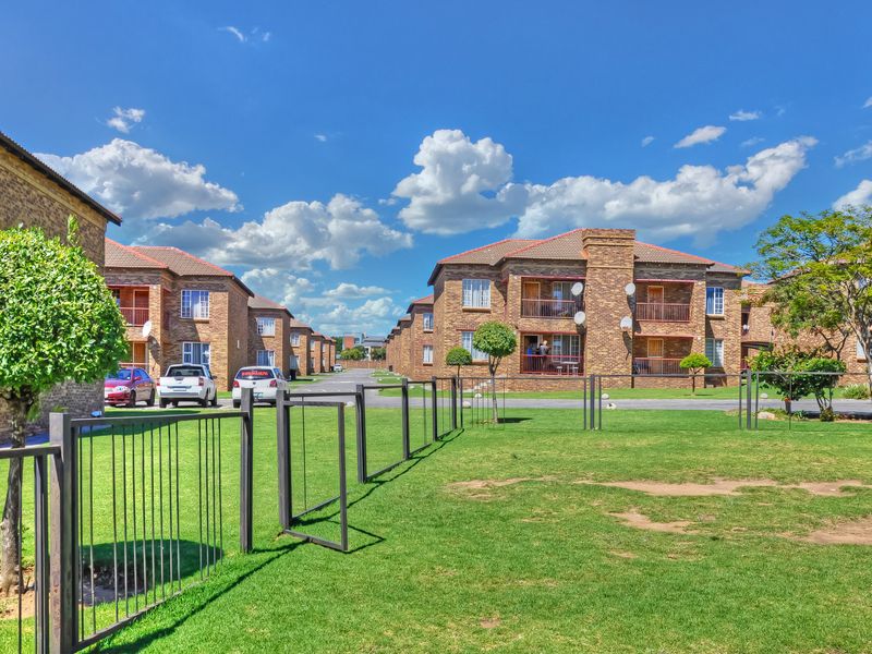 Exterior view of a multi-story apartment complex featuring face brick construction, tiled roofs, and communal garden areas with metal perimeter fencing.