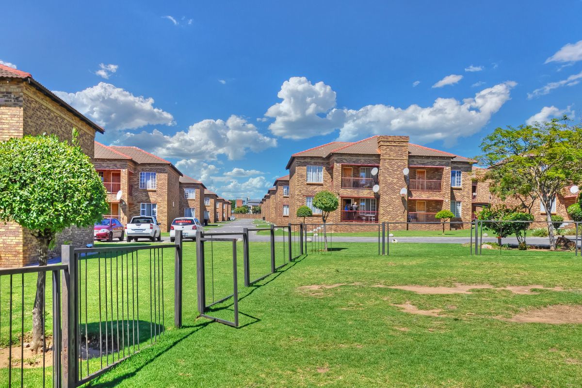 Exterior view of a multi-story apartment complex featuring face brick construction, tiled roofs, and communal garden areas with metal perimeter fencing.