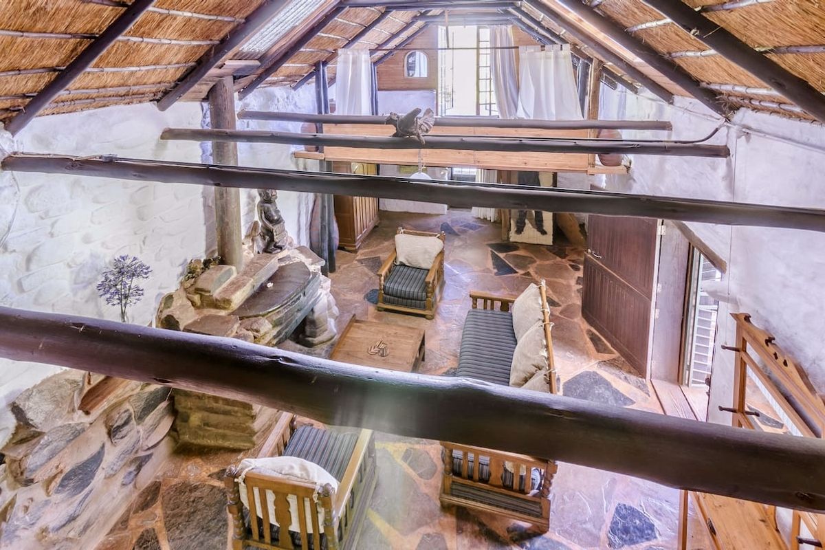Spacious living area featuring a rustic thatched roof, exposed timber beams, flagstone flooring, and a prominent stone fireplace with a mezzanine level above.