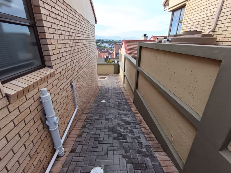 A narrow paved side passage between townhouse units featuring brick walls, a boundary wall with a water tank, and distant neighborhood views.