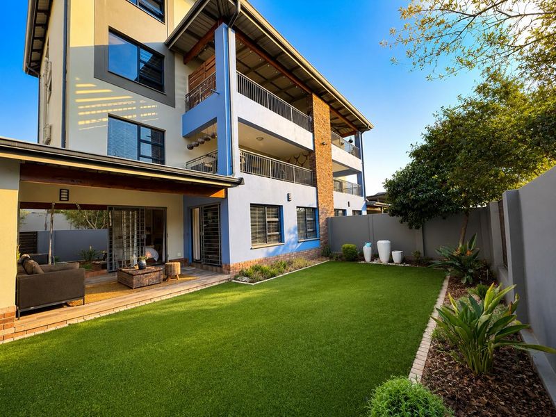 A well-maintained rear exterior view of an apartment building featuring a covered patio, private garden with artificial lawn, and multiple balconies.