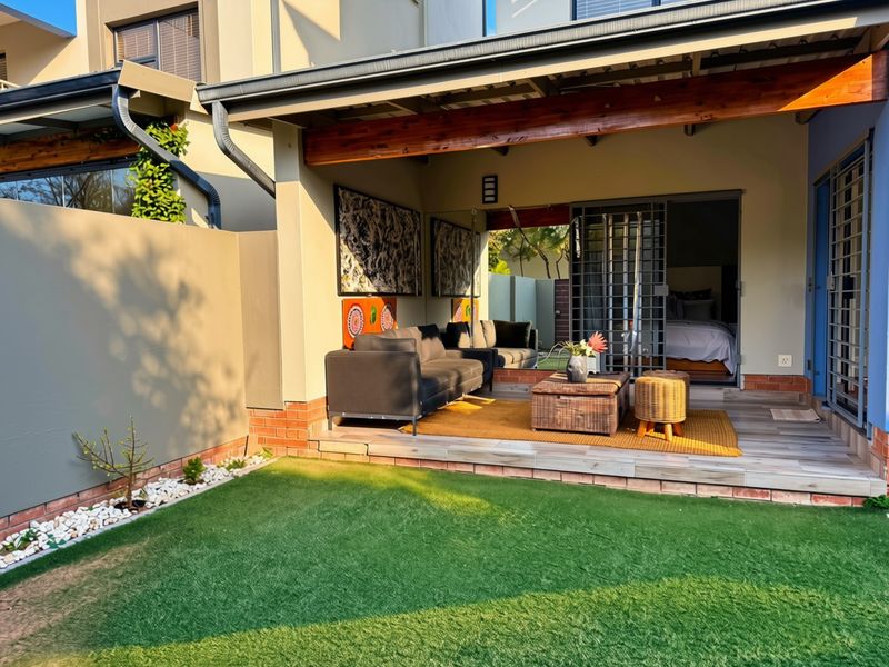 A covered patio area featuring tiled flooring, a timber beam, and a security gate, leading out to a small garden with artificial lawn.