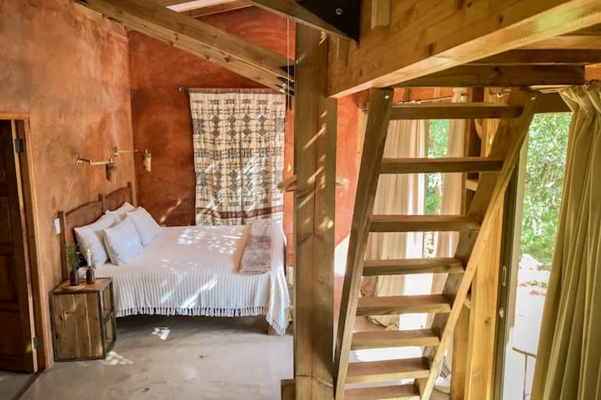 A rustic bedroom featuring exposed timber rafters, a mezzanine staircase, polished concrete floors, and warm textured walls with natural light.