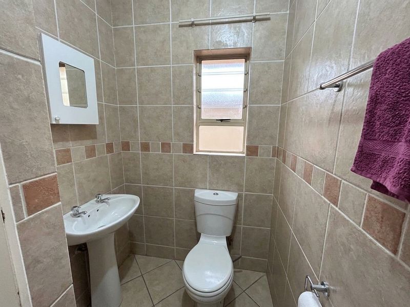 A compact bathroom featuring neutral floor-to-ceiling wall tiles, a pedestal basin, and a standard toilet with a louvered window for ventilation.