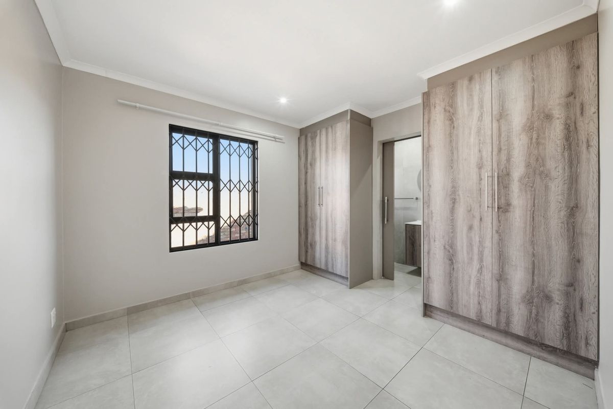 A bright, modern bedroom featuring neutral tiled flooring, built-in wood-grain cupboards, and a window fitted with security bars.