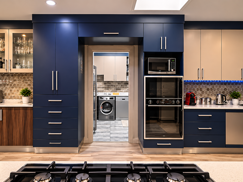 Modern kitchen featuring high-contrast navy and wood cabinetry, stone countertops, hexagonal tile backsplash, and premium built-in appliances with a view into the laundry area.