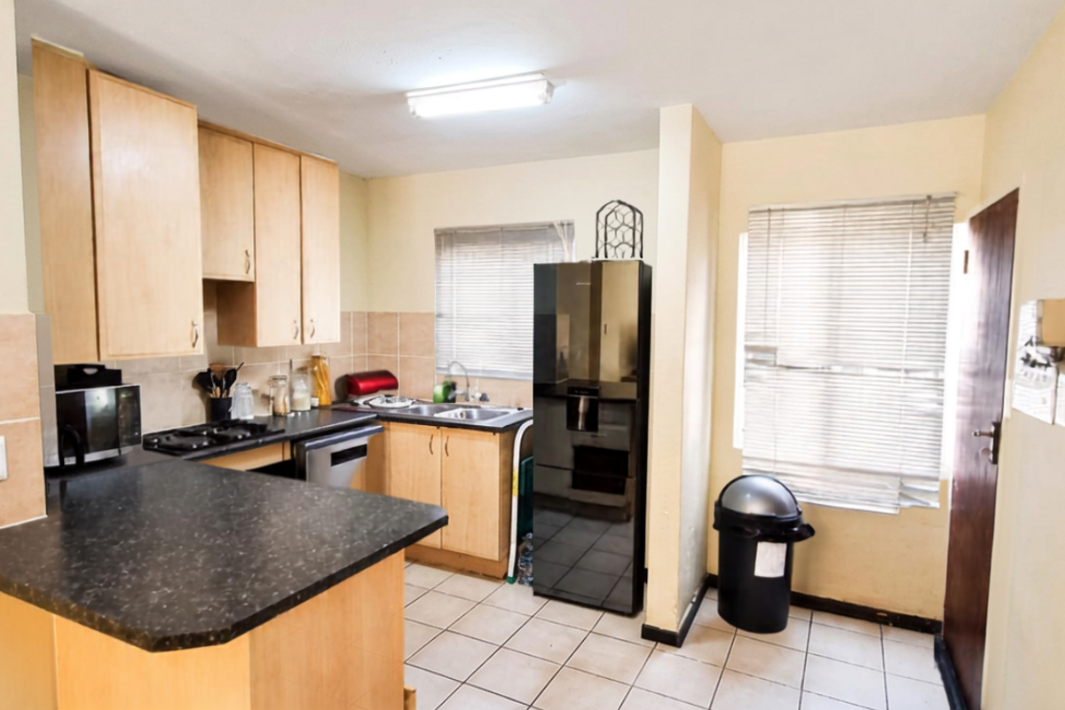 A functional kitchen featuring laminate cabinetry, dark countertops, ceramic tile flooring, and a double bowl sink with natural light from window blinds.
