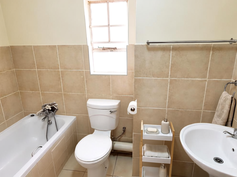 A functional bathroom featuring a bathtub, pedestal basin, and toilet with neutral wall tiling and a window for natural light.