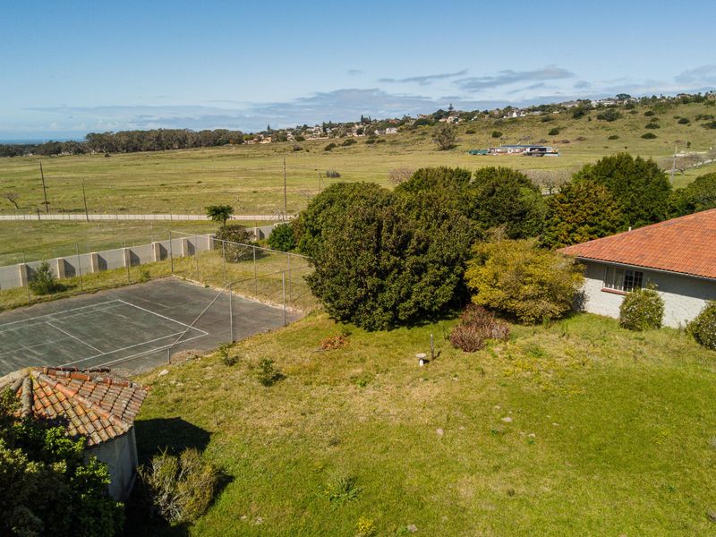 Aerial view of the property grounds featuring a private tennis court, expansive lawn, and surrounding landscape with distant coastal views.