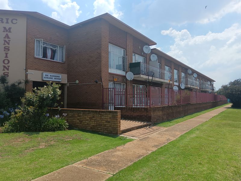Exterior view of Ric Mansions apartment complex featuring brick facade, balconies, and perimeter security fencing along a paved walkway.