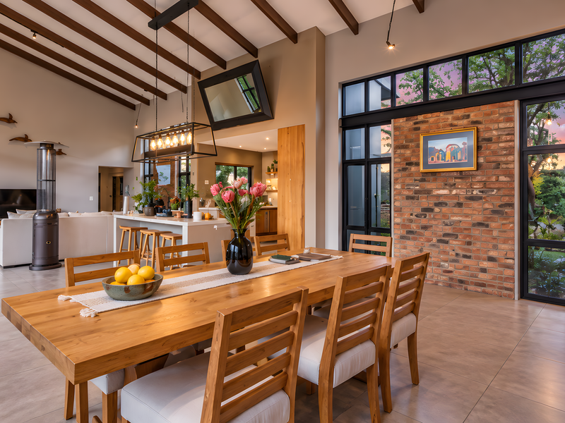 Spacious open-plan living and dining area featuring exposed timber beams, a striking brick feature wall, and large windows overlooking the garden.