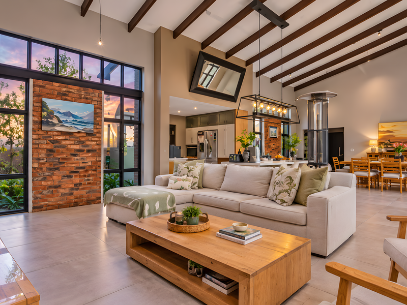 Spacious open-plan living and dining area featuring high vaulted ceilings with exposed timber beams, large windows, and a striking exposed brick feature wall.