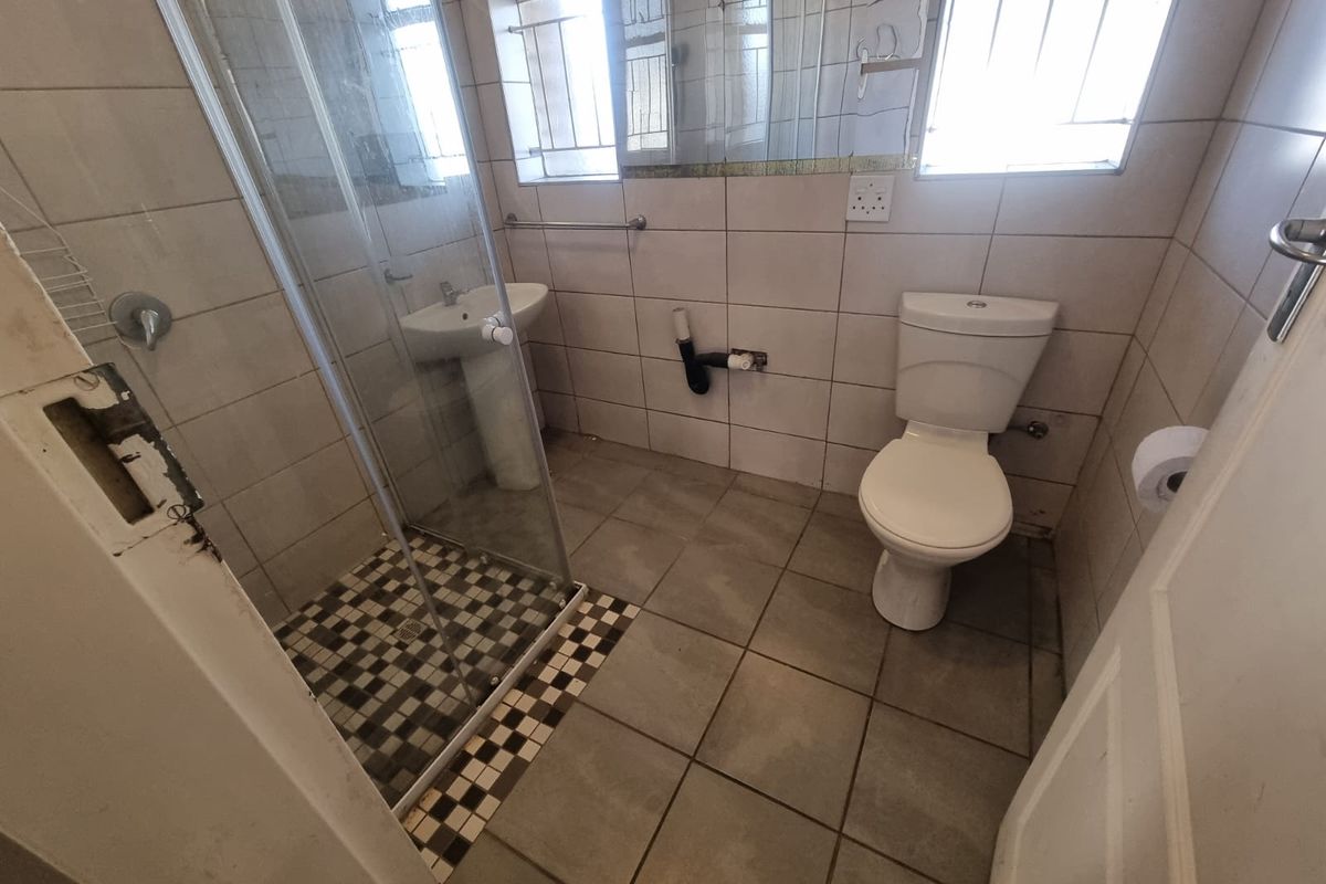 A functional bathroom featuring a shower enclosure, pedestal basin, and toilet, with neutral wall and floor tiling throughout.