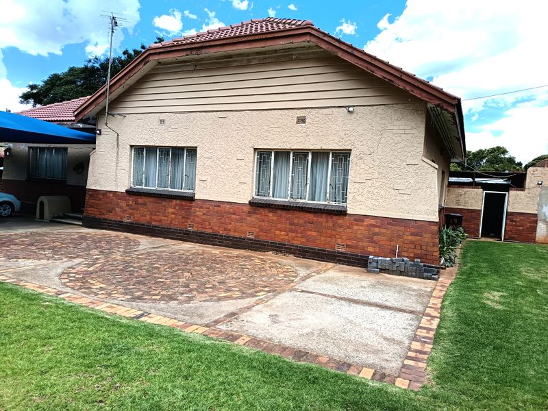 Rear view of a single-story house featuring a brick and plaster facade, tiled roof, and a paved patio area leading to a lawn.