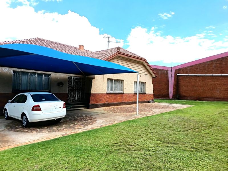 Front exterior view of a single-story house featuring a paved driveway, a blue shade sail, and a well-maintained lawn area.