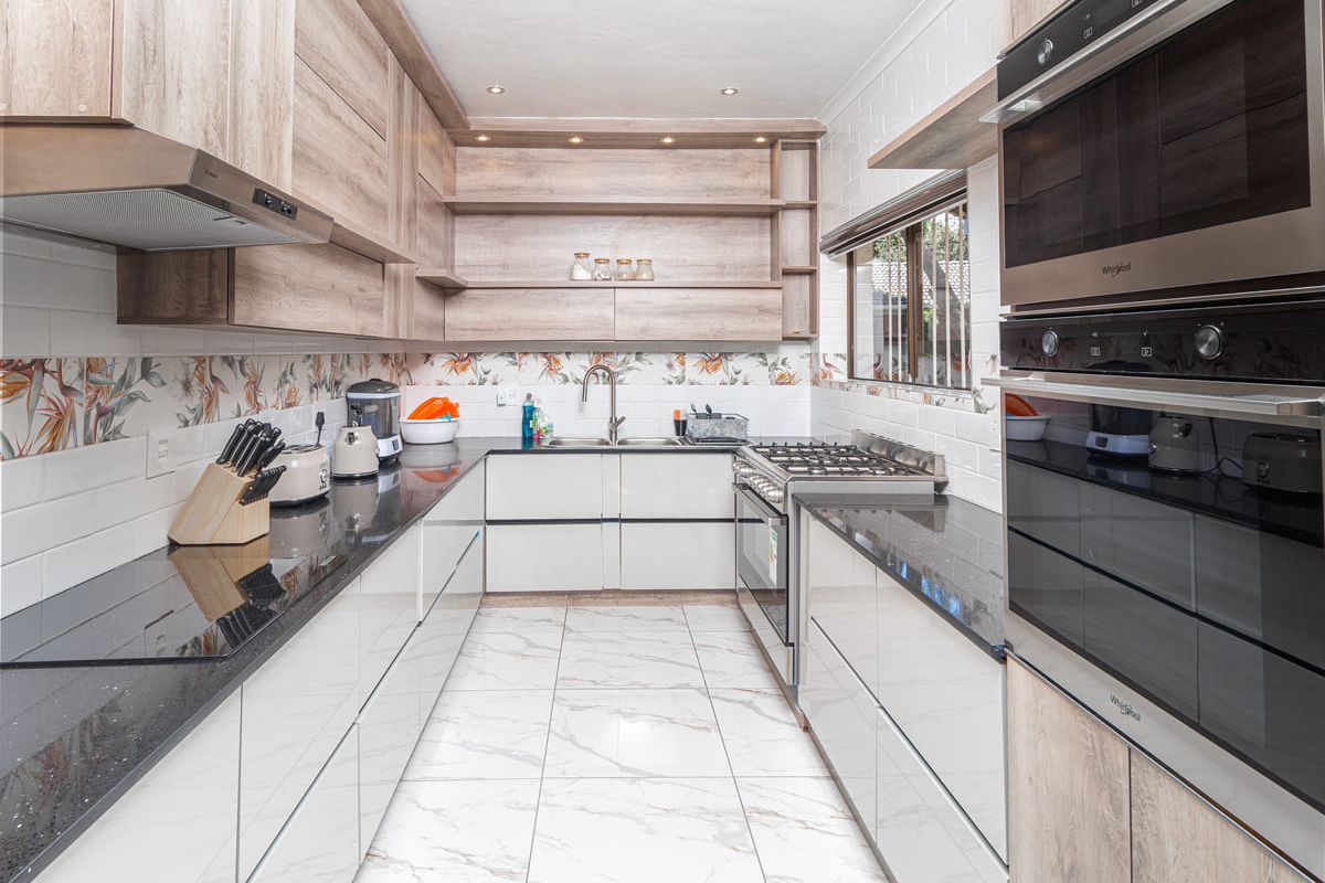 Modern galley-style kitchen featuring sleek white cabinetry, wood-tone open shelving, black granite countertops, and integrated stainless steel appliances including a gas hob and wall oven.