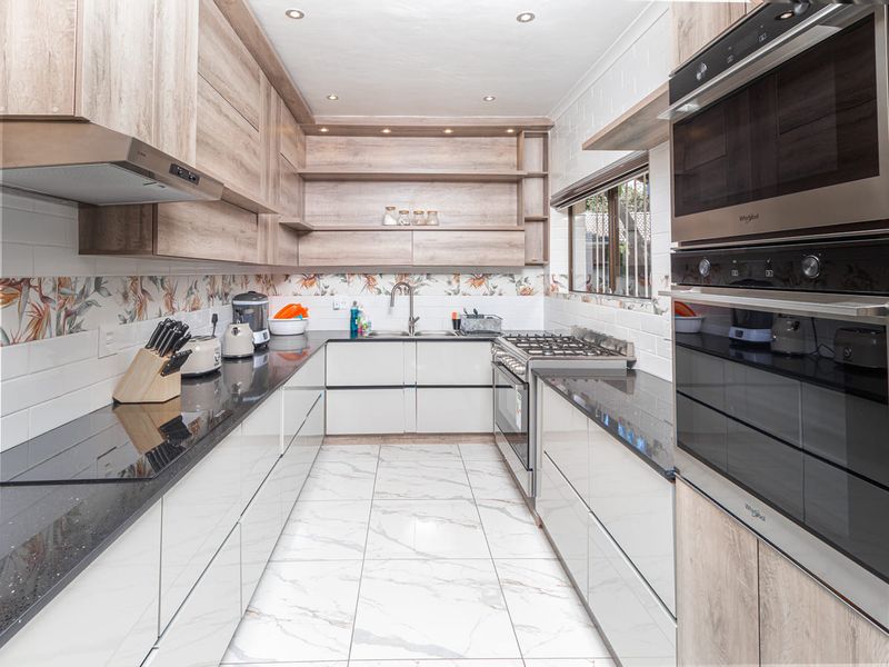 Modern galley-style kitchen featuring sleek white cabinetry, wood-tone open shelving, black granite countertops, and integrated stainless steel appliances including a gas hob and wall oven.